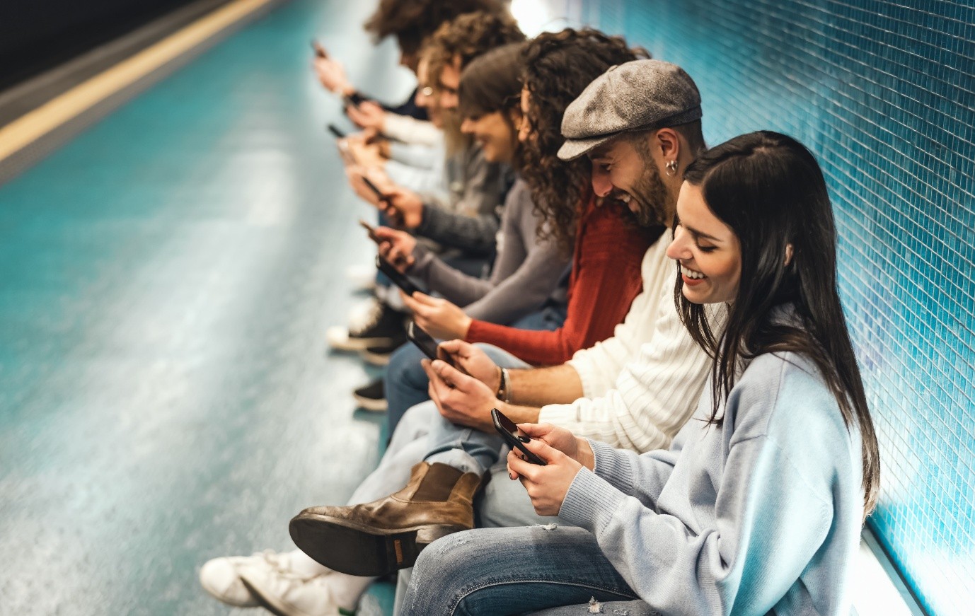 A group of people sitting together in a row on a bench, each focused on their mobile phone screen and smiling.